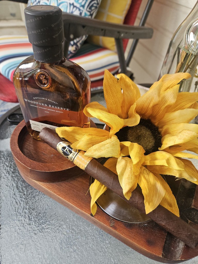 "Cigar and Woodford Reserve Bourbon on a porch beside sunflowers."